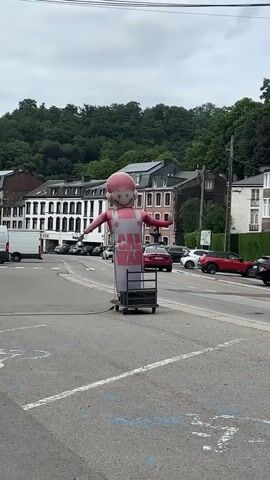 This media file shows an outdoor scene in Spa, Belgium. The primary subject is an inflatable advertising figure, resembling a cartoon character with a pink head and wearing a white apron. The apron has red text that reads "CAR WASH". The inflatable is positioned on a cart with wheels in the middle of what appears to be a street or parking lot.

In the background, there are several parked cars and a white van. Further back, buildings line a street that curves uphill. These buildings have a European architectural style, with tiled roofs and multiple stories. A lush, green hillside covered in trees rises behind the buildings.

The weather appears to be overcast, with a cloudy sky. The time of day seems to be daytime, likely late morning or afternoon, given the overall lighting. The setting is a public area, possibly a town street or commercial district, given the presence of businesses and traffic.

The activity depicted is essentially a static advertisement, with the inflatable figure standing in place. There are no people actively interacting with the inflatable or the immediate surroundings in the frame. The overall mood is neutral, focusing on the visual elements of the scene and the advertisement.