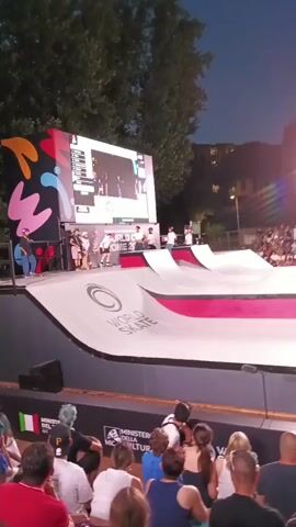 This is a vertical, full-shot video of a nighttime skateboarding event. The camera is positioned behind a seated audience, looking towards a large, illuminated skate ramp. In the background, a large screen displays an image of skateboarders, and beyond that, stadium lights illuminate a crowded spectator area. The skate ramp itself is white with red accents and features a "World Skate" logo. Several people, some in athletic gear, are on the ramp, and one person is actively skateboarding. The audience in the foreground is partially visible, with many people facing the ramp. The overall atmosphere appears to be that of a sporting competition.