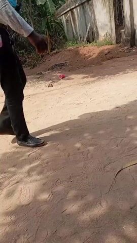 The video shows a man walking on a dirt path towards a concrete wall. The man is wearing a light-colored button-down shirt, black pants, and black shoes. A small pink fan is attached to his belt. As the man walks away, two women in colorful skirts and blouses walk in the opposite direction, carrying purses. The background includes trees, a concrete wall topped with barbed wire, and a red plastic cup on the ground. The dirt path is covered in footprints.