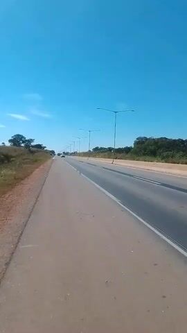 A first-person perspective video captures a view of a paved road with a clear blue sky above. The road has multiple lanes, and streetlights are visible along the right side. There is sparse vegetation and trees on either side of the road. Several vehicles are driving on the road. The camera appears to be moving along the road.