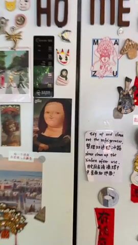 A refrigerator door opens, revealing its contents. On the exterior of the door, several magnets are affixed, including a caricature of the Mona Lisa, a clown-like figure, and a small, green-and-pink striped doll. A printed note with both English and Chinese writing is also attached. Inside the refrigerator, shelves are stocked with various food items. On the top shelf, jars of condiments and yellow containers are visible. The second shelf holds a yellow basket, bags of produce, and what appears to be a watermelon. The third shelf contains a metal bowl and more bags of produce, including lemons and green beans. The bottom shelves show additional produce and clear containers. The interior light of the refrigerator illuminates the stored items.