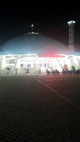 The video shows a mosque at night. The mosque has a large, dome-shaped roof. There is a minaret next to the mosque. The mosque is well-lit. There are several motorcycles parked in front of the mosque. The parking area is paved with dark-colored bricks.