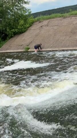 A man, wearing a dark top and dark shorts, is kneeling and reaching out over the edge of a concrete dam into a fast-flowing river. The water is churning and foamy. The dam has a grassy slope behind it, and there are trees to the left. The sky is partly cloudy.