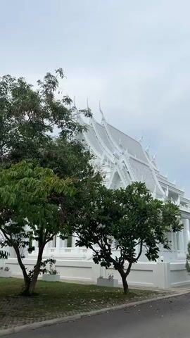 The video shows the exterior of a white Thai temple or religious building. The building has a traditional Thai architectural style, with a multi-tiered, gabled roof and ornate decorations.  There are several trees in front of the building, partially obscuring the view. The trees are green and leafy, and the grass is green and well-maintained. The sky is overcast and gray, suggesting that it may be a cloudy or rainy day. The camera pans slightly, revealing more trees and a paved area. There are no people visible in the video.