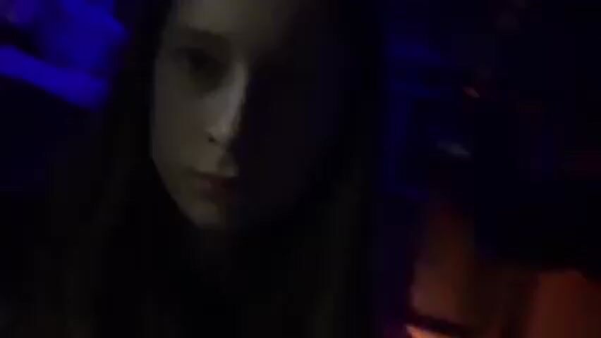 A young woman with long, straight, light brown hair is the primary subject of the video. She is looking directly at the camera with a neutral expression. The environment is dimly lit, with some areas illuminated by blue light. Behind her is a hallway with a dark and slightly industrial appearance, featuring what appears to be a metal structure with a series of narrow openings. The lighting is uneven, and shadows are prominent, making it difficult to discern further details about the surroundings. The camera appears to be hand-held and relatively stable.