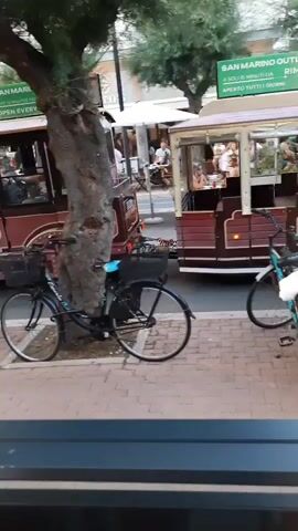 A tree is in the foreground, its trunk thick and textured. Two bicycles are parked beside the tree, one black and one white with red accents. Behind the bicycles, a train with several carriages is passing by. Passengers are visible inside the carriages, some sitting and some standing. The train is decorated with wooden panels and has an open-air design, allowing for views of the interior. A banner with text and images is visible above the train. The ground is paved with bricks. In the background, buildings and more trees can be seen. The overall impression is of a sunny outdoor scene, likely in a town or resort area.