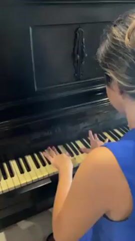 A woman from the back is playing a black upright piano. She is wearing a blue top. The piano has white and yellow keys and an ornate design in the center.