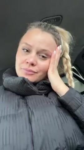 A woman with blonde hair in a ponytail braid is sitting in a car wearing a black puffer jacket. Her left hand is resting on her cheek. She is looking at the camera with a slightly bored expression. Someone is speaking in Russian in the background.
