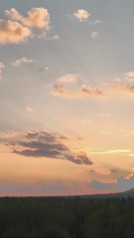 A time-lapse video shows a sunset over a forest and mountains. The sky is filled with clouds that are lit by the setting sun, creating rays of light. The sun is partially obscured by clouds and mountains. The forest is dark and silhouetted against the bright sky. There are no people or animals visible in the video.
