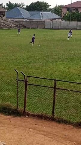 The video shows a soccer match in progress on a green grass field. Players in white and purple uniforms are actively engaged in the game, running and moving the ball. The video is shot from behind a chain-link fence, and a few spectators can be seen in the background. The weather appears to be rainy. Buildings and vegetation surround the field, which is bordered by the fence. The camera's view provides a clear view of the players' actions and the layout of the soccer field.