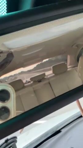 The video is captured from inside a car, looking out the rear window and reflected in the rearview mirror. The interior of the car features tan leather seats. Outside, a road with a slight curve is visible. In the distance, there are hills and trees. At one point, a flock of sheep can be seen grazing near the road. The sky suggests it is either sunrise or sunset, with warm, golden light. A phone is visible in the foreground, reflecting in the mirror.