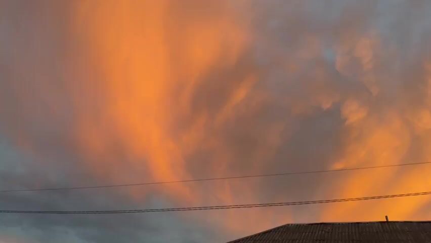 The video shows a time-lapse of a sunset. The sky is filled with dramatic clouds, painted in hues of orange, pink, and gray. The sun is setting in the background, casting a warm glow across the clouds. In the foreground, the silhouette of a roofline and power lines are visible, suggesting an outdoor or semi-urban setting. The clouds are dynamic, shifting and changing shape as the sun descends. The overall impression is one of natural beauty and the transient nature of time.
