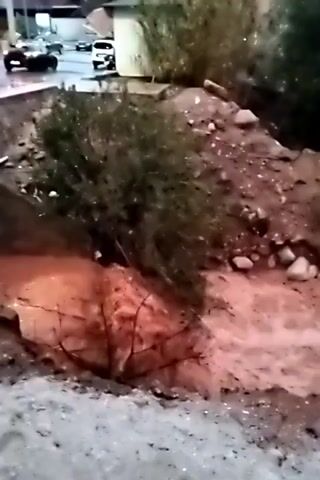 The video shows a muddy stream, likely a flooded drainage ditch or similar channel, coursing through an area of reddish-brown soil and low vegetation. The water appears murky and discolored, suggesting it is carrying a significant amount of sediment. In the background, a landscape of hills and trees is visible under an overcast sky, contributing to an overall impression of a wet or post-rain environment. In the far distance, there is a glimpse of what appear to be cars and buildings, indicating the scene is situated in or near a populated area. The camera is held at a somewhat unsteady angle, panning across the scene, focusing on the flow of the water. The person filming is saying something in another language, but the audio is unclear enough that the language cannot be determined.