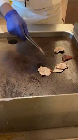 A close-up shot shows a chef wearing blue gloves and a white chef's coat as they use metal tongs to flip slices of meat on a flat-top grill. The grill is dark and appears to have some residue on it. Several slices of meat are already on the grill, some of which are browning. In the background, a yellow container and part of a buffet station are visible. The lighting is bright, suggesting an indoor setting, likely a kitchen or restaurant.