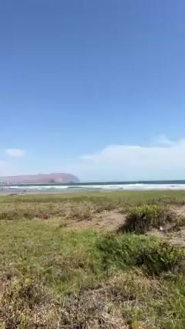 The video shows a coastal scene on a bright, sunny day. A sandy and grassy area stretches towards the ocean, with gentle waves breaking on the shore. The sky is mostly clear with a few wispy clouds. A distant landmass is visible on the horizon. As the video progresses, it pans slightly, revealing a sign placed in the grassy area. The overall atmosphere is peaceful and natural.