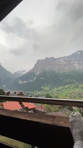 This media file captures a serene moment with a breathtaking mountainous backdrop. The scene opens with a panoramic view of lush green valleys dotted with small houses, leading up to a majestic, snow-capped mountain range under a cloudy sky. The perspective suggests the viewpoint is from a balcony or terrace, with a silver railing in the foreground. Two pairs of feet, clad in grey socks, are visible, suggesting relaxation.

The scene then shifts to focus on two individuals. A young woman with long, dark hair and a headband is smiling at the camera. Beside her, a young man wearing a blue t-shirt and a baseball cap is also looking towards the camera. They appear to be sitting on a balcony or patio, with wooden structures and windows visible behind them. The overall mood is one of comfort and contentment.

The location cues from the majestic mountains and the village architecture strongly suggest Grindelwald, Switzerland. The cloudy weather implies it could be morning or late afternoon. No visible text is present in the media file.
