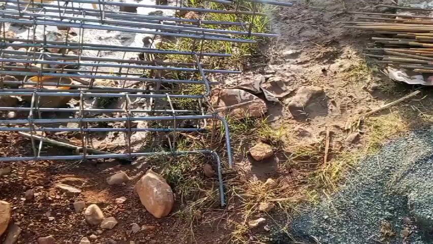 A construction site is captured in a close-up shot. The focal point is a metal rebar cage laid on the ground, surrounded by dirt, rocks, and sparse patches of grass. The rebar forms a grid pattern with both horizontal and vertical members, and it is partially elevated off the ground by bent legs at its corners. The surrounding environment includes a mix of soil types, from dark brown to reddish-brown, with small and medium-sized rocks scattered around. In the background, glimpses of a blue roof and green foliage suggest a building under construction or renovation. The scene conveys an early stage of construction, specifically foundation work involving reinforcing the concrete with rebar.