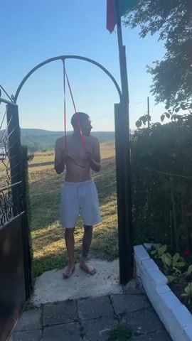 The video begins with a close-up of a young man standing in an outdoor setting, shirtless and wearing light-colored shorts. He appears to be holding a red ribbon or rope, with a pole and an arched structure behind him. The background shows a grassy hill and trees, suggesting a rural or semi-rural location. The lighting indicates it is daytime, likely late afternoon or early evening, given the warm sunlight.

The scene then shifts to a garden area. A white-painted raised flower bed is filled with various colorful flowers, including white daisies, pink roses, and purple blossoms. Lush green foliage surrounds the flowers, and a tall tree with sunlight filtering through its leaves is visible in the background. A wire fence runs along the back of the garden. In the far distance, the silhouette of a vehicle can be seen. The bright sunlight creates a lens flare effect in some shots, enhancing the warm and sunny atmosphere. The overall impression is of a peaceful, warm day in a garden, possibly in a village or countryside setting like Bogdanovo, Bulgaria, as suggested by the location context. No text is visible in the video.