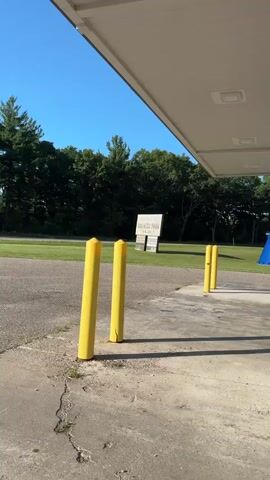 The video shows a gas station on a sunny day. The camera pans across several gas pumps and a blue silo. A red fire extinguisher is mounted on a post. In the background, there are trees and a blue sky. The video also includes a shot from inside a car looking out the window at the gas station. There are no people visible in the video.
