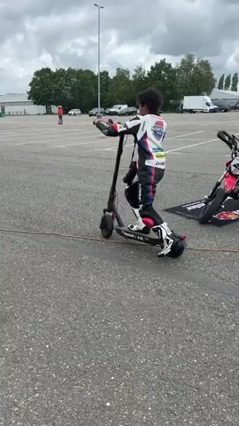 A person in a white and black racing suit is riding a black electric scooter on an asphalt surface. They are wearing a helmet and appear to be practicing maneuvers. In the background, there are trees, buildings, cars, and orange cones set up on the ground, suggesting an outdoor training area or parking lot. The sky is overcast. Another person on a motorcycle is visible in the distance, also participating in activities.