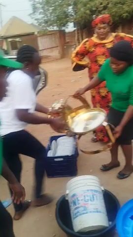 The video shows a group of people outdoors. In the foreground, three women are handling serving dishes and a blue crate filled with white plates. One woman is wearing a green shirt and black skirt and holding golden serving stands. Another woman is in a white shirt and black pants. A third woman stands in the background wearing a colorful orange and red outfit with a head wrap. Nearby are a white bucket and a large black tub with a blue lid.

To the right, a man dressed in a white shirt is looking at his phone. Behind him is an orange drink cooler on a green stool. The environment seems to be a yard or outdoor area with a building and a car visible in the background. The ground appears to be dirt.