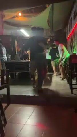 The video shows a brightly lit outdoor bar area at night. The bar has a covered roof structure with various decorative lights. Several people are present, including a couple who are dancing in the center of the space. Other patrons are seated at the bar or on stools, some talking and drinking. A bartender is visible behind the counter. The flooring appears to be made of concrete or tiled material. The atmosphere suggests a casual social gathering.