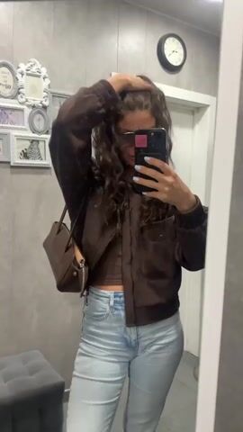 A woman is taking a mirror selfie indoors. She has long, wavy brown hair, is wearing dark sunglasses, and has light blue nail polish on her fingers. She is dressed in a dark brown cropped jacket, a matching dark brown crop top, light blue high-waisted jeans, and dark brown shoes. She holds a black smartphone with a pink sticker on the case, and a dark brown handbag hangs from her left shoulder.

The setting appears to be a room with light grey, textured walls. In the background, there's a white door and a wall adorned with several ornate, empty picture frames in various sizes and shades of grey, with one frame visibly containing a zebra print. A round black clock showing approximately 10:15 is mounted on the wall above the door. To the left, a grey upholstered ottoman or stool is partially visible. The woman adjusts her hair briefly at the beginning of the video, then poses for the camera while looking at her phone. A white-framed mirror reflects the scene.