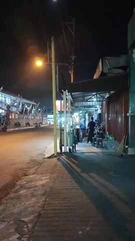 The video is a vertical, low-light shot taken at night from a sidewalk looking towards a street and a row of businesses. A streetlamp illuminates the left side of the frame, casting long shadows across the sidewalk and street. On the sidewalk, a yellow pole with electrical wires runs vertically through the center of the frame. To the right of the pole, a green cart is visible. In the background, several people are standing and sitting in front of a business with a dark facade. Their faces are not clearly visible. On the left side of the street, a long building with banners is visible, and several motorcycles are parked in front of it. The overall impression is of a quiet urban street scene at night.