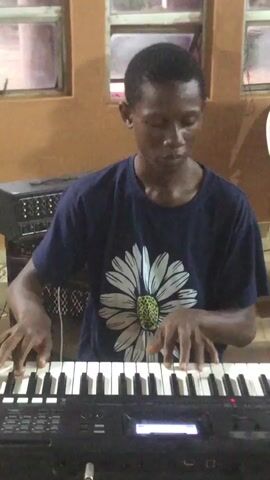 In this media file, a young Black male is seated in front of a keyboard. He is wearing a dark blue t-shirt with a large white daisy graphic on it. His hands are positioned on the keys, and he appears to be playing the instrument. The keyboard has a digital display screen and several buttons and knobs.

Behind the individual, the setting appears to be indoors. The walls are a warm, earthy orange color. There are windows with visible panes in the background, suggesting a building. To the left of the individual, a piece of audio equipment with a rack of knobs and buttons is visible. To the right, a white plastic chair is partially visible. The lighting suggests it is daytime, but there are no clear indicators of the specific time or weather conditions. The overall impression is that of a casual setting, possibly a home or a community space where music is practiced or performed. The individual's facial expression is neutral, focused on the task of playing the keyboard.