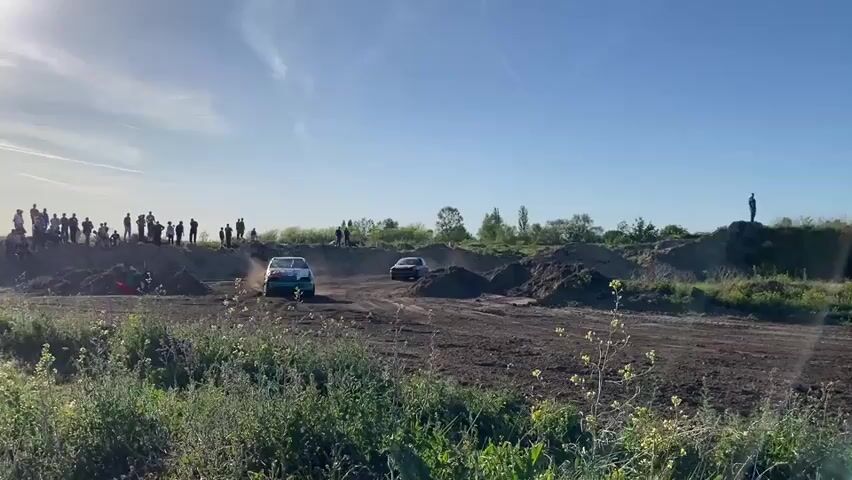 This video captures an autocross or rallycross event taking place on a sunny day in Kanjiza Municipality, Serbia. The scene depicts a dusty, unpaved track surrounded by dirt mounds and green vegetation, including bushes and wildflowers in the foreground.

Two racing cars are central to the action, navigating a sharp turn and creating a substantial cloud of reddish-brown dust. The lead car, a dark-colored coupe, prominently displays the number "201" and the text "3 MAJ" on its side as it powers through the corner. The second, lighter-colored car is almost entirely engulfed by the dust cloud generated by the lead vehicle.

Spectators are visible observing the race from various vantage points. A crowd is gathered on a raised dirt bank to the left of the track, while another individual stands atop a higher dirt mound on the right, silhouetted against the clear blue sky. Distant trees frame the horizon under the bright, clear conditions. The considerable amount of dust indicates dry weather and the high-speed nature of the motorsport event, creating an atmosphere of dynamic competition.