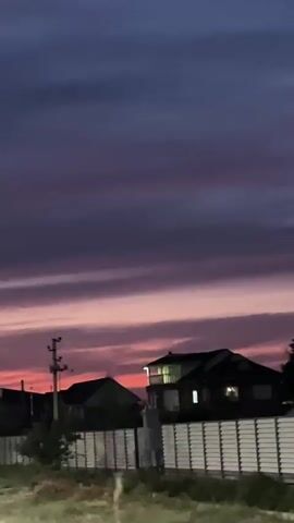 The video shows a blurry image of a suburban landscape at dusk. The sky is a mix of deep purple and pink hues, with horizontal bands of clouds. In the foreground, there is a white fence with horizontal slats. Behind the fence, there are several houses with lights on in the windows. There are also some trees and a utility pole visible. The camera appears to be moving slightly, causing some of the image to be out of focus.