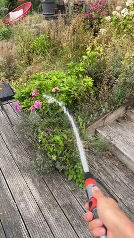 A hand holding a red and gray hose nozzle sprays water onto a garden bed. The garden bed contains various plants, including green bushes, pink and purple flowers, and dry, brown grasses. Wooden planks form a deck in the foreground, and a brick wall is visible in the background. There are also planters with plants and a red chair in the background. The scene appears to be outdoors on a sunny day.