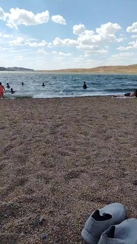 The video shows a beach with people swimming in the water. The beach is covered in small pebbles. In the foreground, there is a pair of white shoes. In the background, there are hills and a cloudy sky. The water is choppy and there are waves. Some people are standing in the water, while others are swimming. The overall impression is one of a casual summer day at the beach.