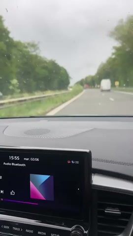 The video captures a first-person perspective from inside a car driving on a road or highway. The car's dashboard and infotainment system are prominent in the foreground. The infotainment screen displays "15:55 mar. 02/06" (Tuesday, June 2nd) and indicates "Audio Bluetooth" is selected, though it currently shows "Aucun titre," "Aucun interprète," and "Aucun album." Buttons for car controls such as "SEEK," "TRACK>," "RADIO," "MEDIA," and "SETUP" are visible below the screen.

Through the windshield, a multi-lane asphalt road is visible, extending into the distance. Other vehicles, including a white van and a red car, can be seen ahead. The sky is overcast and grey, and there are small water droplets or marks on the windshield, suggesting recent rain or spray. Lush green trees and vegetation line both sides of the road. The setting depicts a daytime drive on a highway, likely in or near Enghien, Belgium.