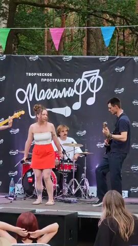 A woman with dark hair pulled back, wearing a white strapless top and an orange skirt, stands center stage, holding a microphone and singing. She has tattoos on her left leg. To her right, a man with dark hair, wearing a dark blue t-shirt and dark pants, plays an electric guitar. Behind them, a man with light brown hair, wearing a white t-shirt, plays a drum kit. The stage has a black backdrop with white lettering and musical notes. Colorful triangular flags are strung above the backdrop. Trees are visible behind the stage. The lighting suggests it is daytime. In the foreground, the backs of two audience members' heads are visible.