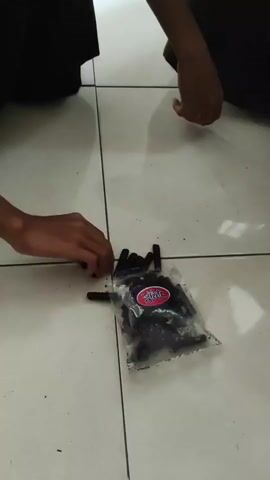 Two individuals are visible, one with their left arm outstretched towards a bag of black cylindrical objects on a tiled floor. The other individual, further away, has their right arm extended towards the bag, also appearing to interact with the objects. The objects are small, black, and appear to be edible, possibly licorice sticks or candy. The bag has a red and blue logo. The floor is light-colored tile. The individuals are wearing dark clothing.