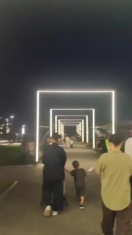 The video begins with a view of a nighttime scene featuring a pathway lined with illuminated rectangular frames. A person pushes a stroller while a child walks alongside, holding their hand. Further down the pathway, other individuals are visible, and to the right, there are benches and foliage. The perspective then shifts to a person in a light olive-colored shirt walking away from the camera, revealing a flagpole with a flag at the top in the background. The scene is illuminated by the pathway lights and ambient lighting.