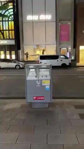 The video shows a Japanese postal box, gray in color, with two slots on top, labeled with Japanese characters and the word "POST." The logo of Japan Post is visible at the bottom of the box. To the left of the box, a person is standing on a ladder, seemingly working on the exterior of a brightly lit storefront. A white van is parked in front of the storefront. The scene appears to be at night.