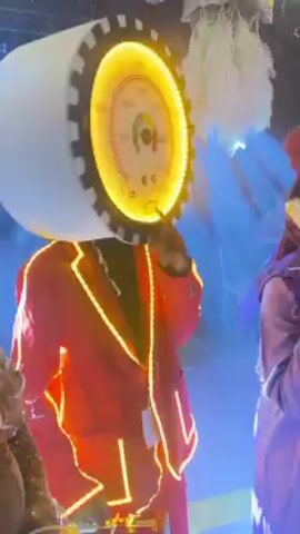 The video shows a person wearing a lit-up costume. The costume consists of a bright orange blazer outlined with LED lights, a briefcase, and a headpiece shaped like a gauge with lights around the rim. The person is dancing on a stage. The lighting is dim and there are spotlights on the stage. There are other people standing in the background.