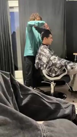 A person with curly hair and wearing a light green jacket and black pants is giving a haircut to a person seated in a barber's chair, draped with a black and white patterned cape. The person receiving the haircut has short, dark hair and is looking to the right. The barber is using scissors and a comb to cut the hair on top of the head. The scene appears to be in a hair salon, with black curtains in the background and a gray couch in the foreground, partially visible. A window is visible behind the curtains.