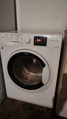This is a video of a washing machine running. The display on the washing machine shows 0:09. The drum of the washing machine is spinning and reflecting light. The washing machine is white and has a black door. It is in a room with a light-colored wall and a wooden floor.