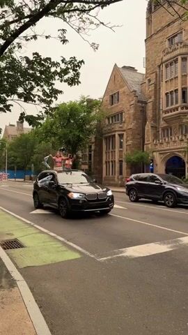 A black BMW X5 drives down a street. Two young women are standing on the roof of the car, waving clothes. Another person is leaning out of a side window, also waving clothes. The car is driving past a large stone building with gothic architecture. In the background, another car is parked. The sky is overcast and hazy.