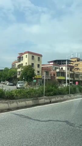 The video shows a view from a vehicle moving along a paved road. On the right side of the road, there is a row of multi-story buildings, mostly residential. The buildings vary in color and architectural style, with some having balconies and air conditioning units visible. There is vegetation lining the road in front of the buildings. The sky above is partly cloudy. The video captures the buildings from a side-angle perspective as the vehicle drives past them. No people are clearly visible in the frame.