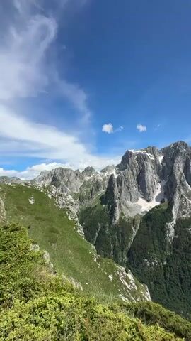 The video shows a panoramic view of a mountainous landscape under a clear blue sky with scattered white clouds. The foreground features green, bushy vegetation. The middle ground consists of rolling green hills and slopes leading towards a range of imposing, rocky mountains. These mountains are rugged and jagged, with patches of snow visible on their higher elevations. Dense green forests cover the lower slopes of the mountains. The scene appears to be a natural, undeveloped environment, likely a national park or wilderness area, captured from a high vantage point.