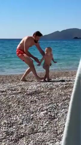 On a sunny day at the beach, a shirtless man wearing orange swim trunks picks up a young child who is wearing a diaper. The man swings the child a couple times and appears to place the child down. The man then picks up the child again and walks along the shore line with the child in his arms. The beach is full of pebbles and there is blue ocean water in the background.