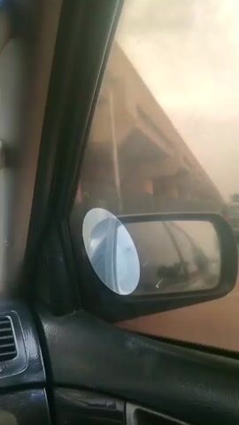 The video appears to be taken from inside a moving vehicle, likely a car, as evidenced by the dashboard and side mirror visible in the frame. The camera moves and shakes, and the view changes as the vehicle drives. The initial shot focuses on the side mirror, then shifts to capture the external environment. Initially, we see an orange three-wheeled vehicle, resembling a tuk-tuk or auto-rickshaw, next to the car. Above it, a man is walking on top of the overhead structure. As the car moves, the camera captures the underside of a bridge or overpass, supported by concrete pillars. Further along, the scene shows a road lined with a curb, some bushes, trees, and street lights, with a figure walking in the distance. The weather appears overcast or cloudy.