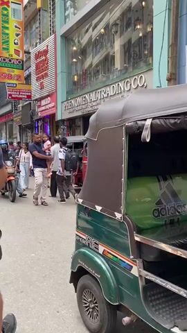The video is a street-level view of a busy urban environment, likely in Asia, with a focus on a street with shops and traffic. The camera is positioned at eye level, moving with a person. In the foreground, a portion of a green and brown three-wheeled auto-rickshaw (tuk-tuk) is visible on the right, with the text "SUPER RE" on its side. The auto-rickshaw appears to be stationary or moving slowly.

On the left side of the street, a person is walking with a large backpack. Another person, a man, is walking towards the camera, also with a backpack. Behind him, a woman with a striped top is walking away. To the left of them, a man is holding a young child. A red auto-rickshaw is in the middle of the street, facing the camera, with a person sitting in the driver's seat. Another red auto-rickshaw is visible further down the street.

The buildings lining the street have multiple stories and feature storefronts. One prominent storefront displays the name "PHENOMENAL TRADING CO." with an address and contact information visible. Another storefront, labeled "U.K.EL," is also visible. In front of the "U.K.EL" store, a small stand is set up with various items, possibly phone accessories.

The street is paved and appears to be a pedestrian walkway and vehicle thoroughfare. Other people are visible walking and operating vehicles, suggesting a lively commercial area. The overall atmosphere is one of active street life and commerce. The lighting suggests it is daytime.
