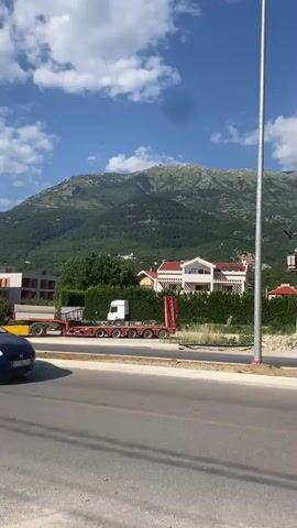 A bright, sunny day with scattered clouds is depicted in this media file, likely taken in Sutvara, Montenegro. The scene is a roadside view, with a paved road in the foreground and a blue car partially visible on the right. In the mid-ground, a white truck is attached to a long, red flatbed trailer carrying what appears to be construction materials or large pipes. Behind the truck and trailer, a well-maintained hedge lines the edge of a property. Further back, several modern houses with red-tiled roofs and white exteriors are nestled amongst lush green trees. Dominating the background is a majestic, green, and rocky mountain range, its slopes covered in dense vegetation. A tall, metallic pole, likely a lamppost or utility pole, stands prominently on the right side of the frame, casting a shadow. The overall impression is one of a peaceful, developing area with a stunning natural backdrop. There is no visible text.