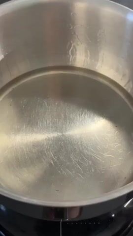 A person's hand with painted fingernails is holding a yellow object, possibly a piece of lemon, over a pot of clear liquid on a stovetop. The hand drops the object into the liquid, and several small, square, yellow pieces, likely food items, are seen frying in the liquid, bubbling and sizzling. The pot is made of stainless steel, and the stovetop appears to be black. The liquid is clear and heated, causing the food items to fry.