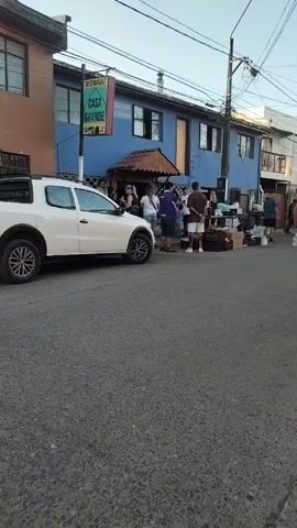 A street scene shows several people standing and walking near a blue building labeled "Restaurant Casa Grande." A white SUV is parked on the left side of the street. Further down the street, people are gathered around what appears to be a street market or yard sale, with boxes and piles of clothes visible. Several young people play with a red ball on the street. An additional child is walking down the street. A baby stroller sits in the foreground on the right. The environment includes buildings, power lines, and a street light. The sky is clear and bright.